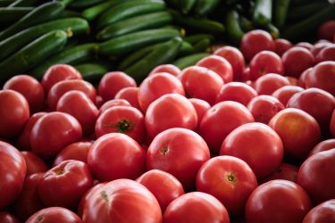 A képen friss paradicsomok láthatók előtérben, mögöttük kígyóuborkák sorakoznak egy piaci standon.