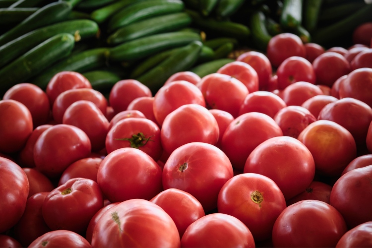 A képen friss paradicsomok láthatók előtérben, mögöttük kígyóuborkák sorakoznak egy piaci standon.