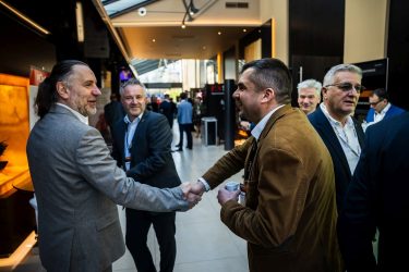 Konferenciahelyszínen üzleti öltözetű résztvevők beszélgetnek, ketten kézfogással üdvözlik egymást, miközben a háttérben további szakmai egyeztetések zajlanak.