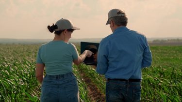 Két gazdálkodó áll egy szántóföldön, és laptopon nézi át a támogatási ügyintézéshez szükséges adatokat.